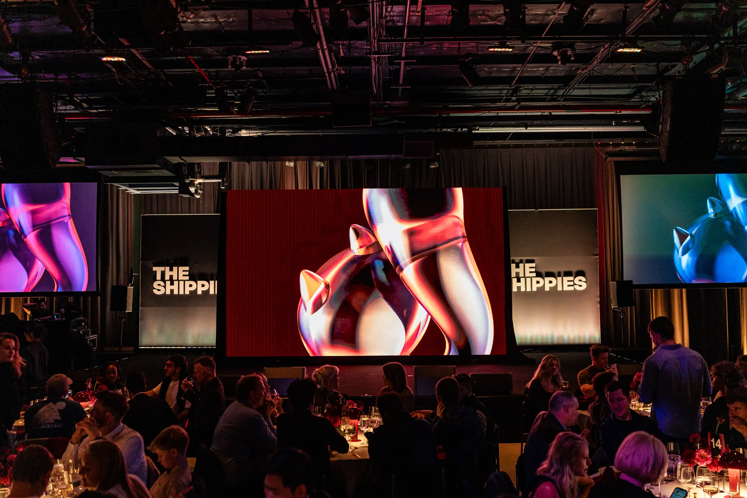 The Shippies ’26 awards night: the trophy animation on three stage screens under the lighting rig, above a dark room of dinner tables